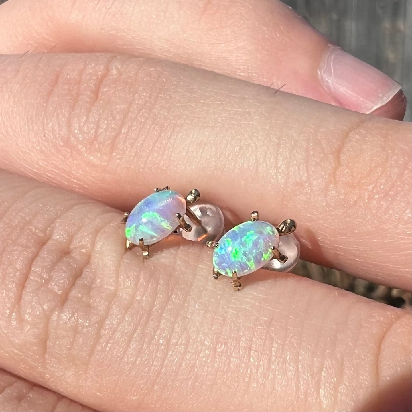 A macro close-up of 14k gold lab-created opal turtle stud earrings in natural sunlight, held at an alternate angle to highlight their deep blue-green opal fire.