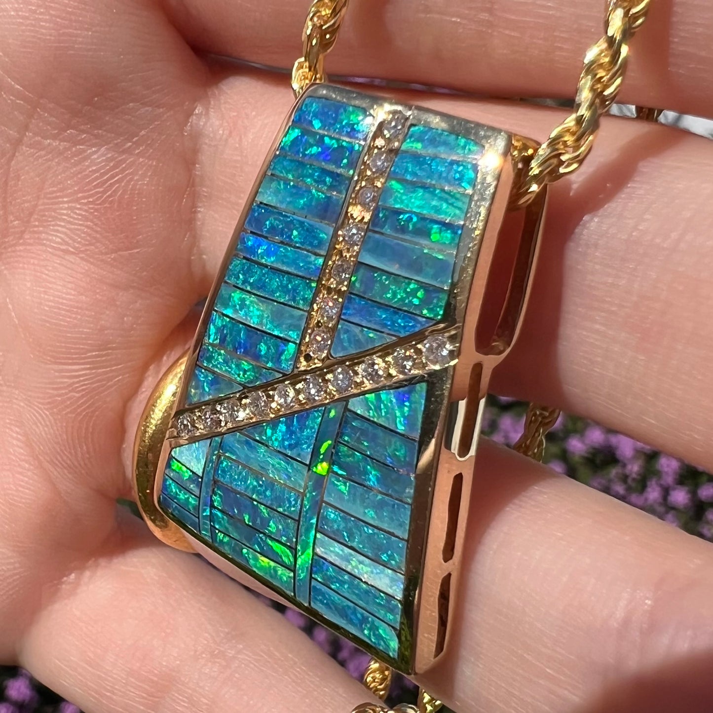 Hand holding an 18k gold opal inlay pendant from an angle in the sunlight. The opals glow with blue and green fire, and diamond accents sparkle.