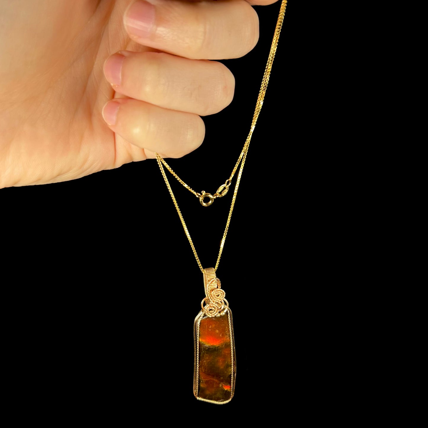 A wire wrapped ammolite necklace held from a hand against a black background.