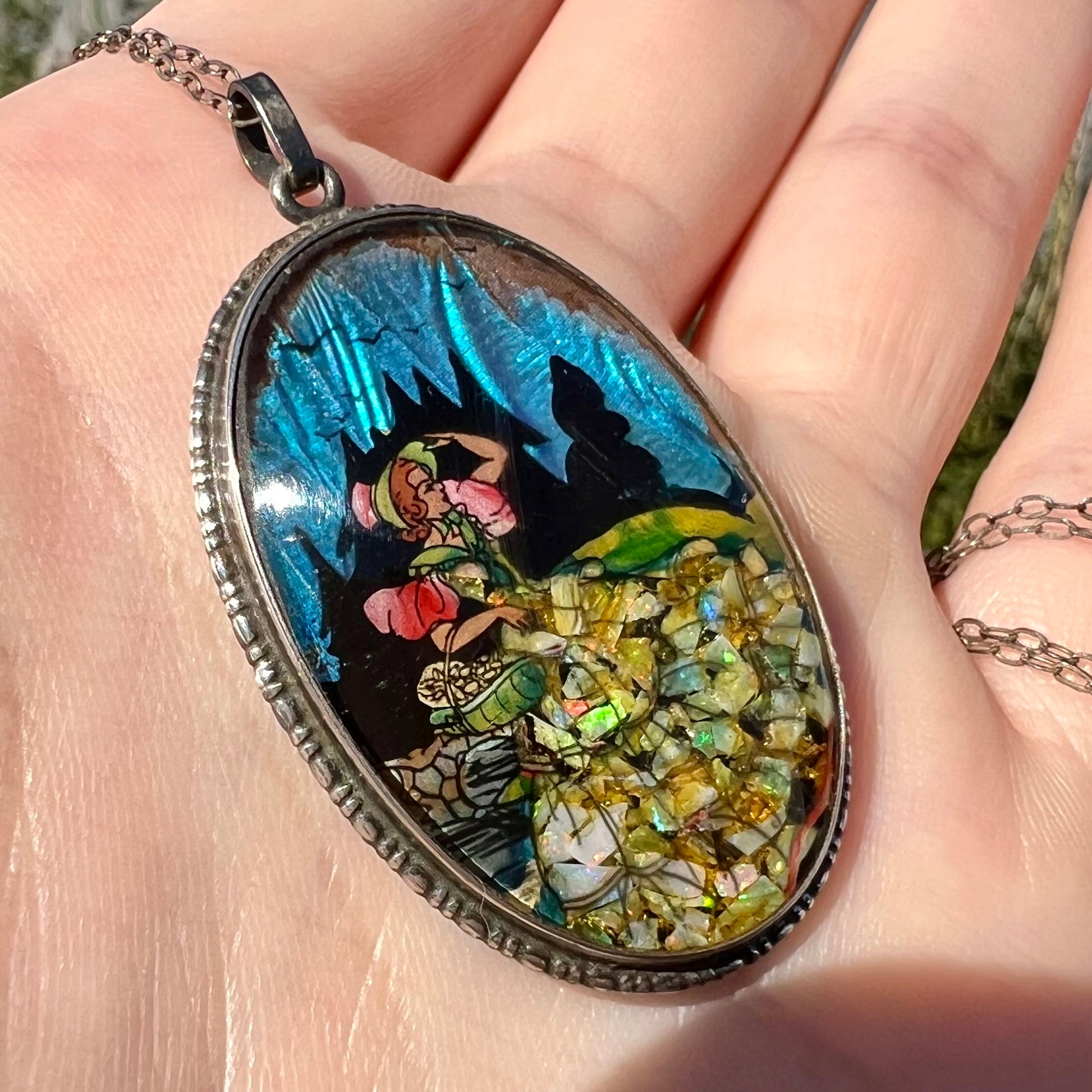 An antique crushed opal and butterfly wing pendant in sterling silver, shown in a hand at an angle in sunlight.