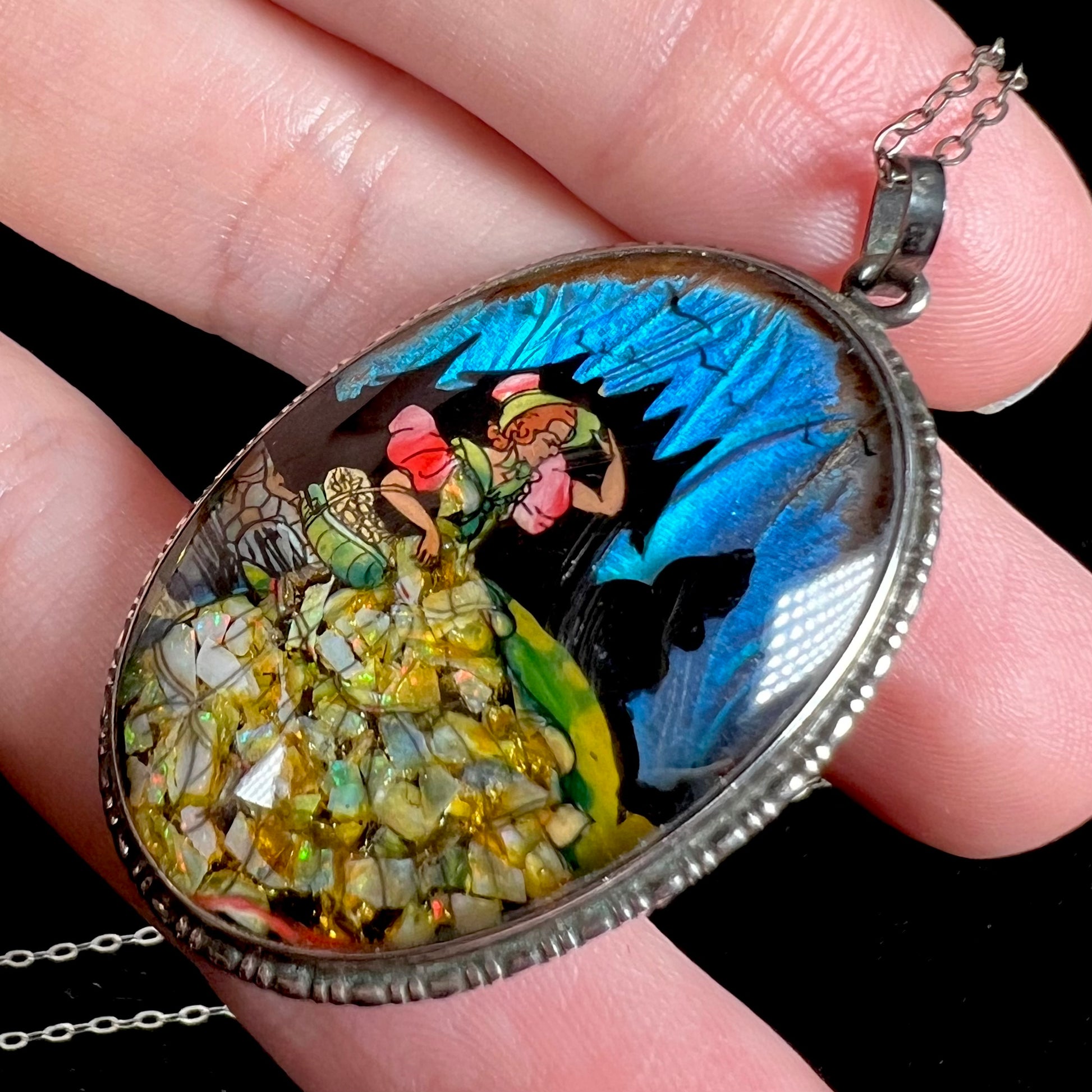 An antique English silver opal and butterfly wing pendant, shown close-up at an angle in a hand against black background.