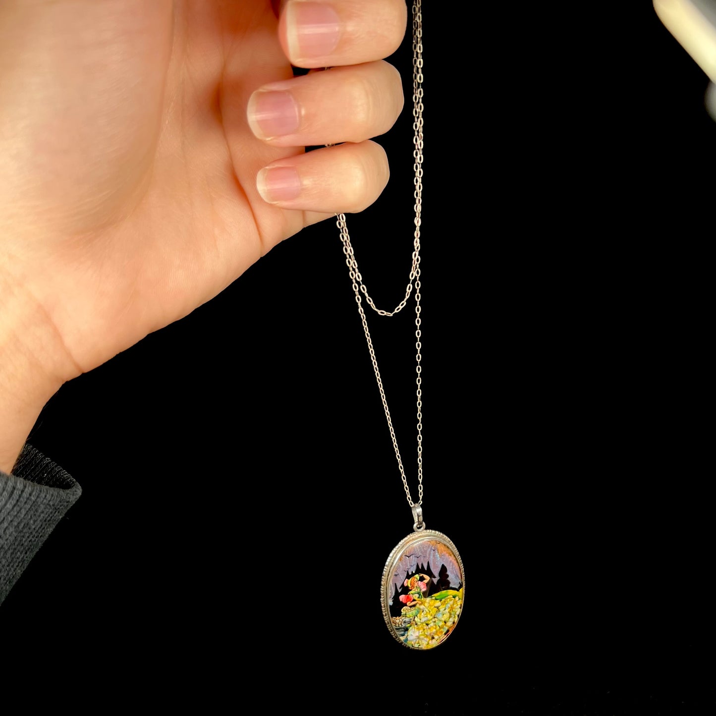 A silver necklace with the image of a woman made from butterfly wing and crushed opal, held by a hand from its chain.