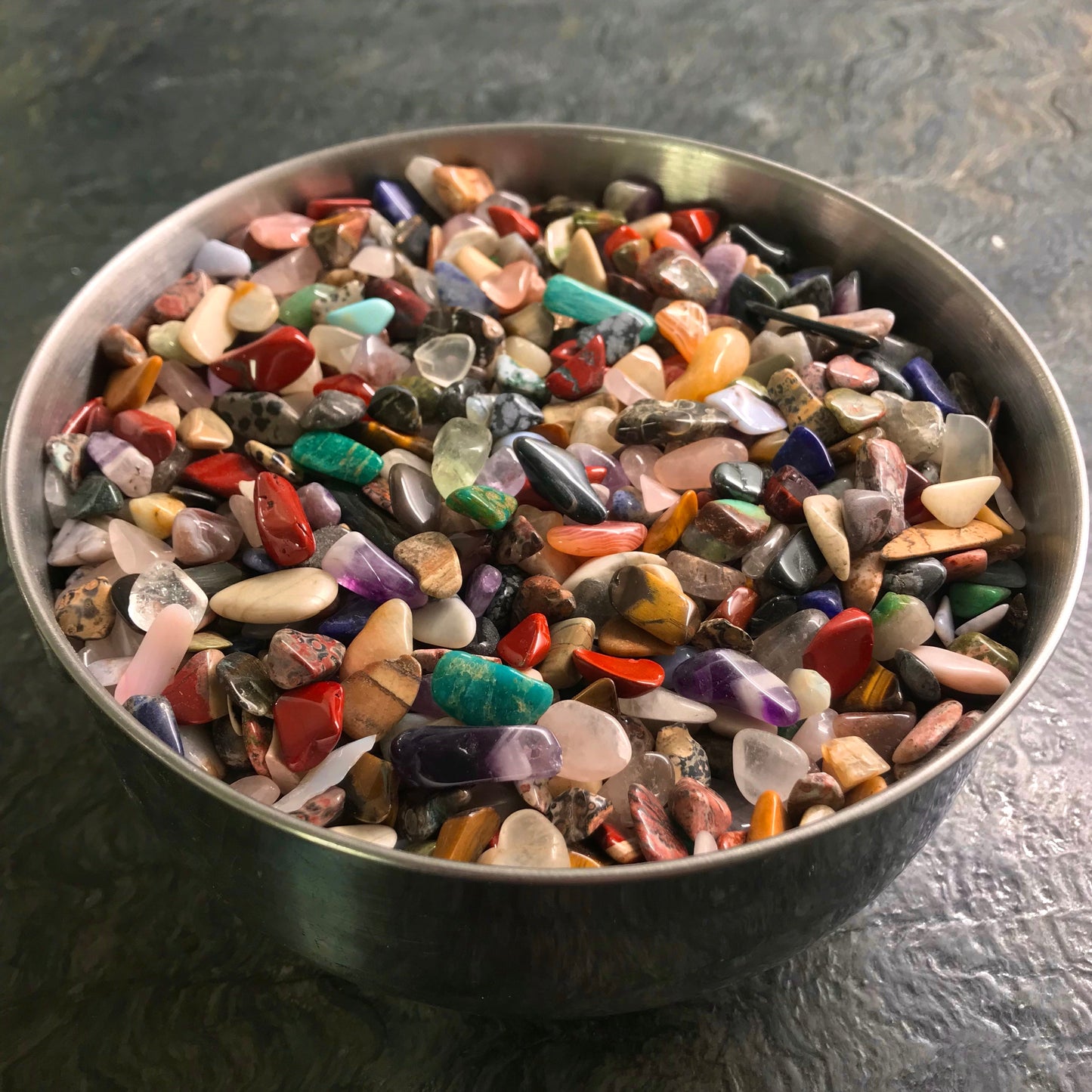 A photo of a round metal bowl on a dark green stone countertop containing hundreds of small tumble polished rainbow gemstones. Red jasper, amazonite, purple amethyst, rose quartz, dalmatian stone, carnelian, sodalite, and more are seen in the bowl. Perfect jewelry making supplies for arts and crafts.
