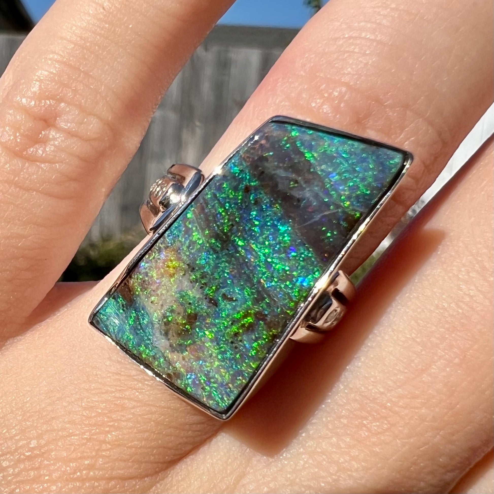 Close-up of a platinum Australian boulder opal ring, worn on a finger in sunlight.  The opal has green color play.