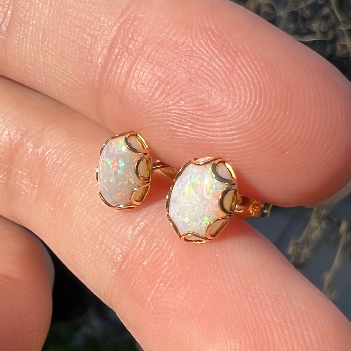 Angled sunlit close-up of a pair of Australian white opal stud earrings in yellow gold held between two fingers.
