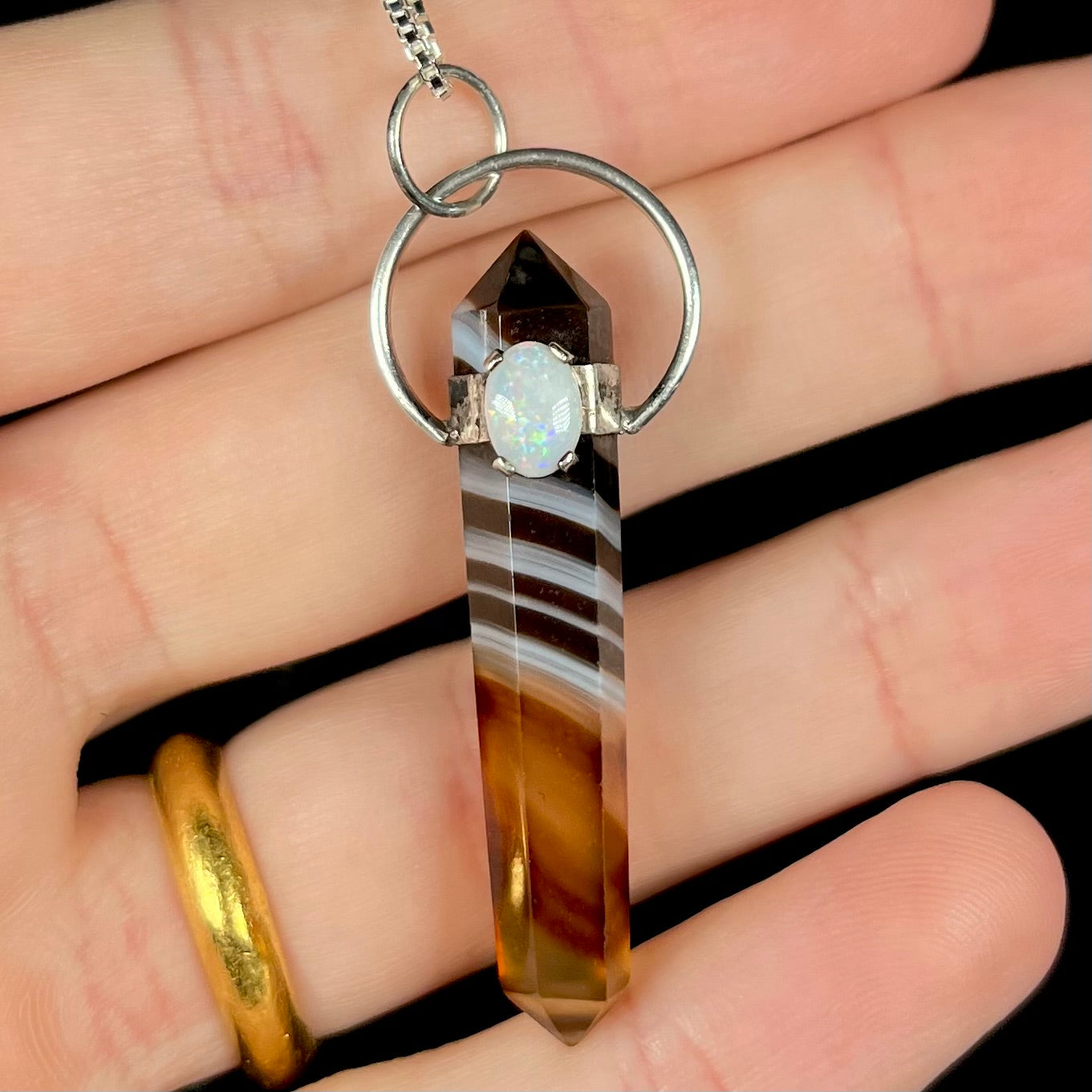 A banded sardonyx opal pendant with an Australian white opal accent, held in fingers against a black backdrop.