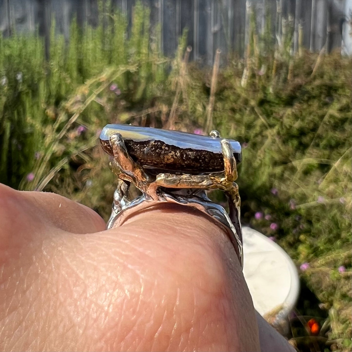 Side gallery profile view of a big boulder opal ring against a blurred outdoor background, worn on a hand in sunlight.