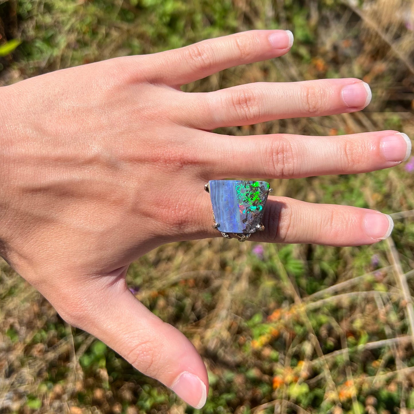 A big boulder opal ring with bright green fire, worn on a hand in the sunlight against a natural backdrop.