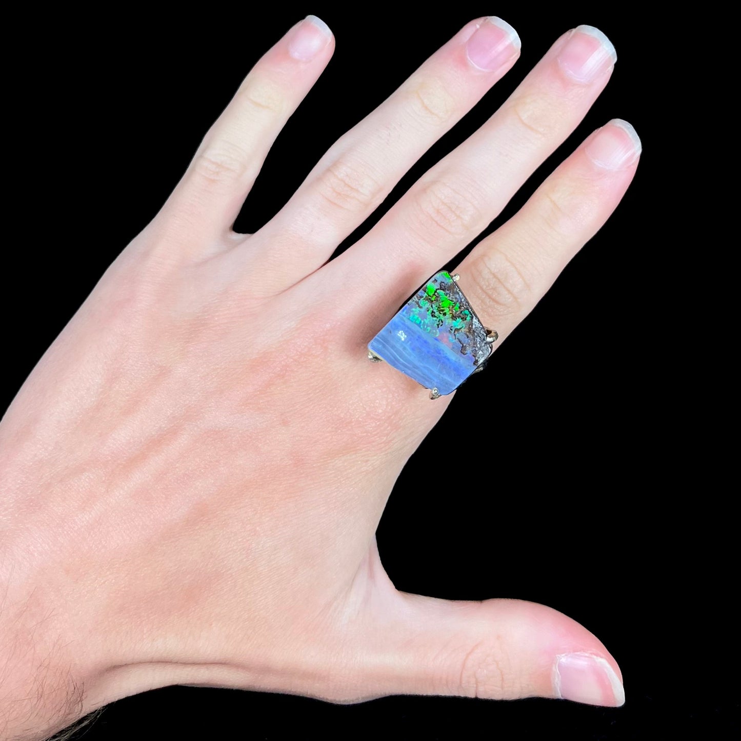 A hand wearing a big boulder opal ring with green flashes seen in the opal's ironstone matrix.