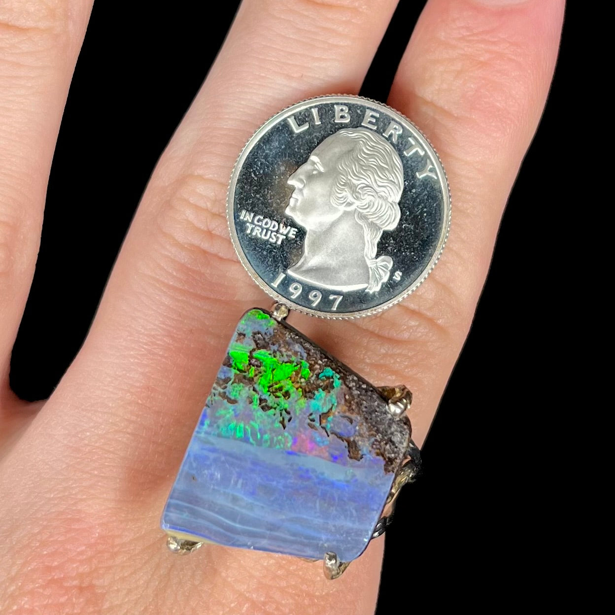 A big boulder opal ring with a bright green flash, worn on a finger next to a quarter coin for scale.