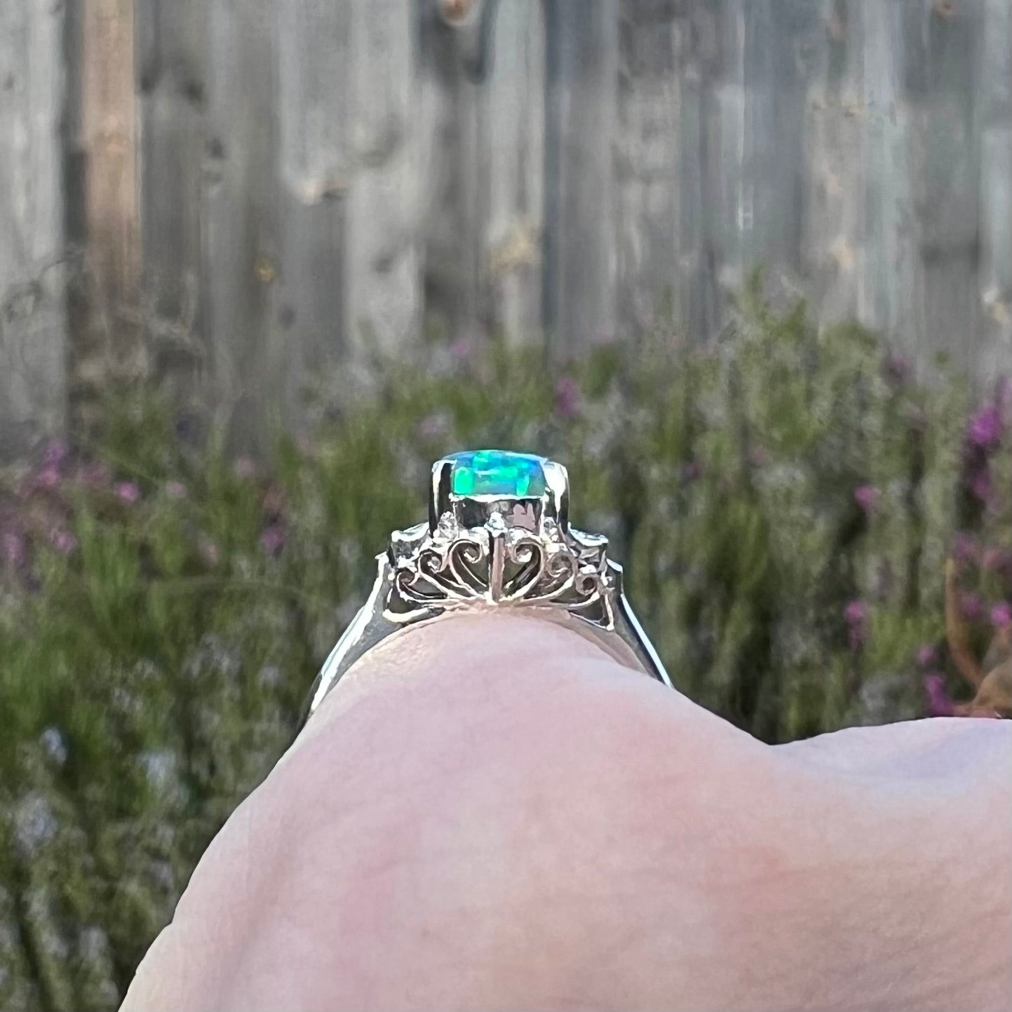 Side gallery view of a platinum black opal and diamond engagement ring, worn atop a pinkie finger on a cloudy day.