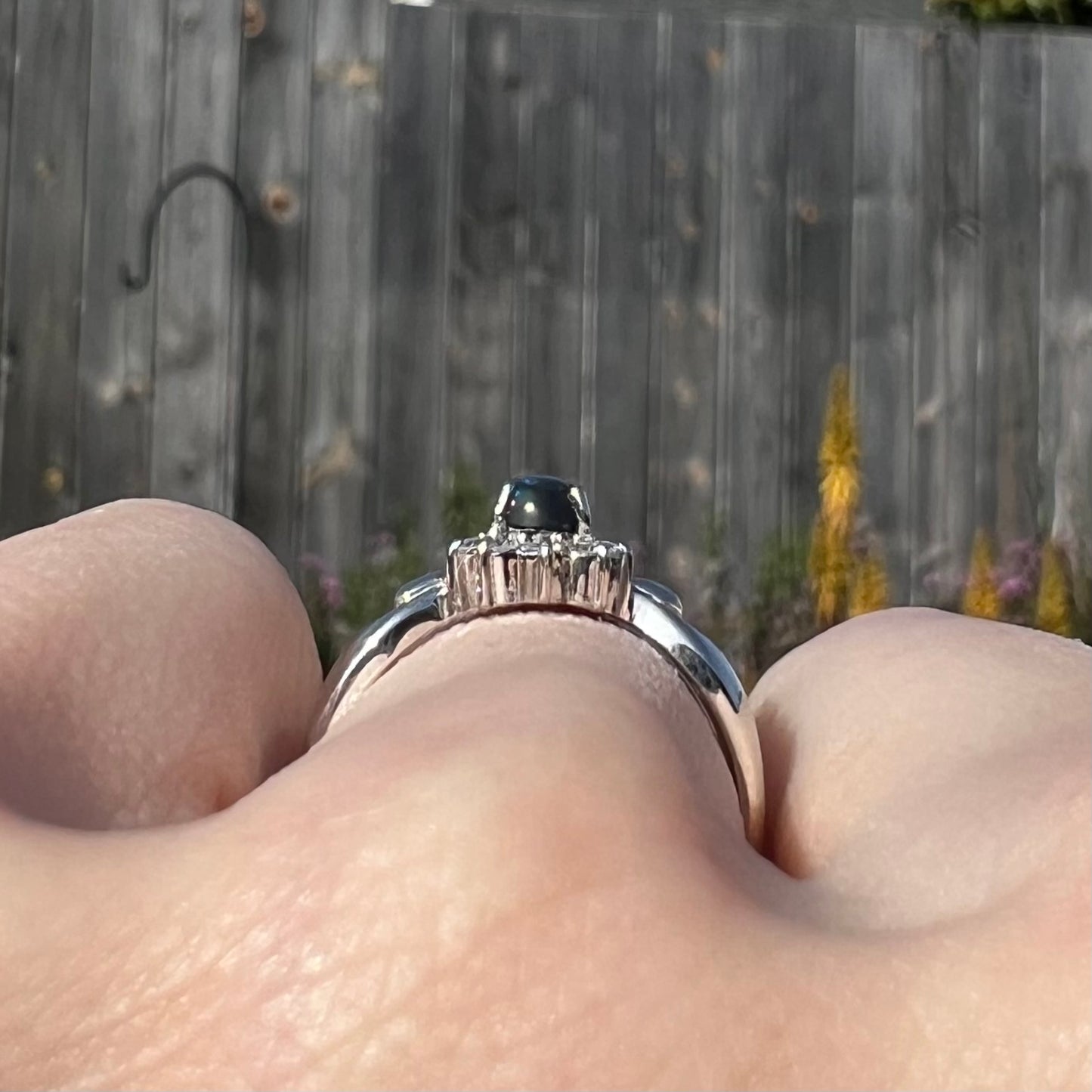 Gallery-style macro view of black opal engagement ring in sunlight with wood fence background.