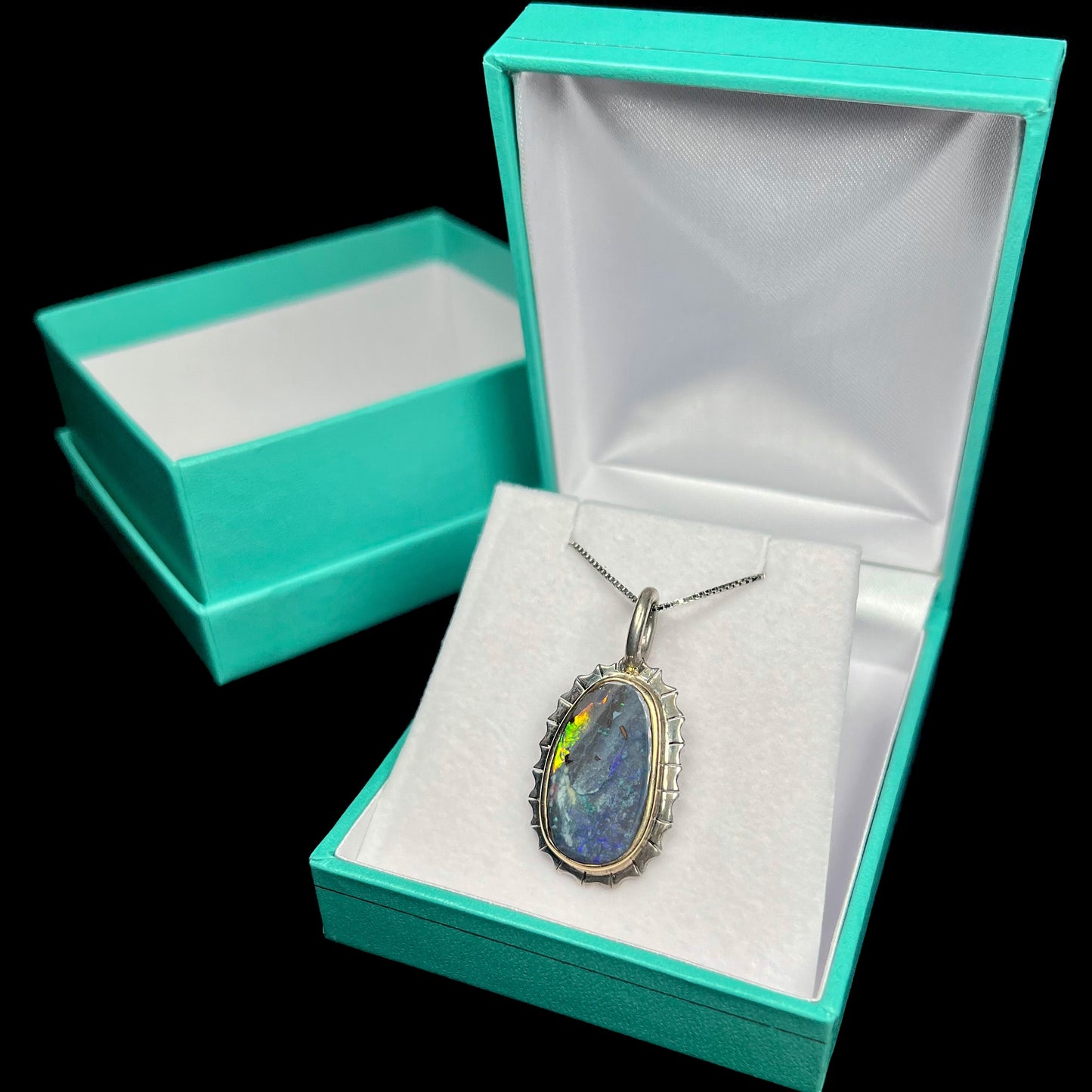 A two-tone silver and gold necklace set with a natural black boulder opal. The necklace is in a blue box.