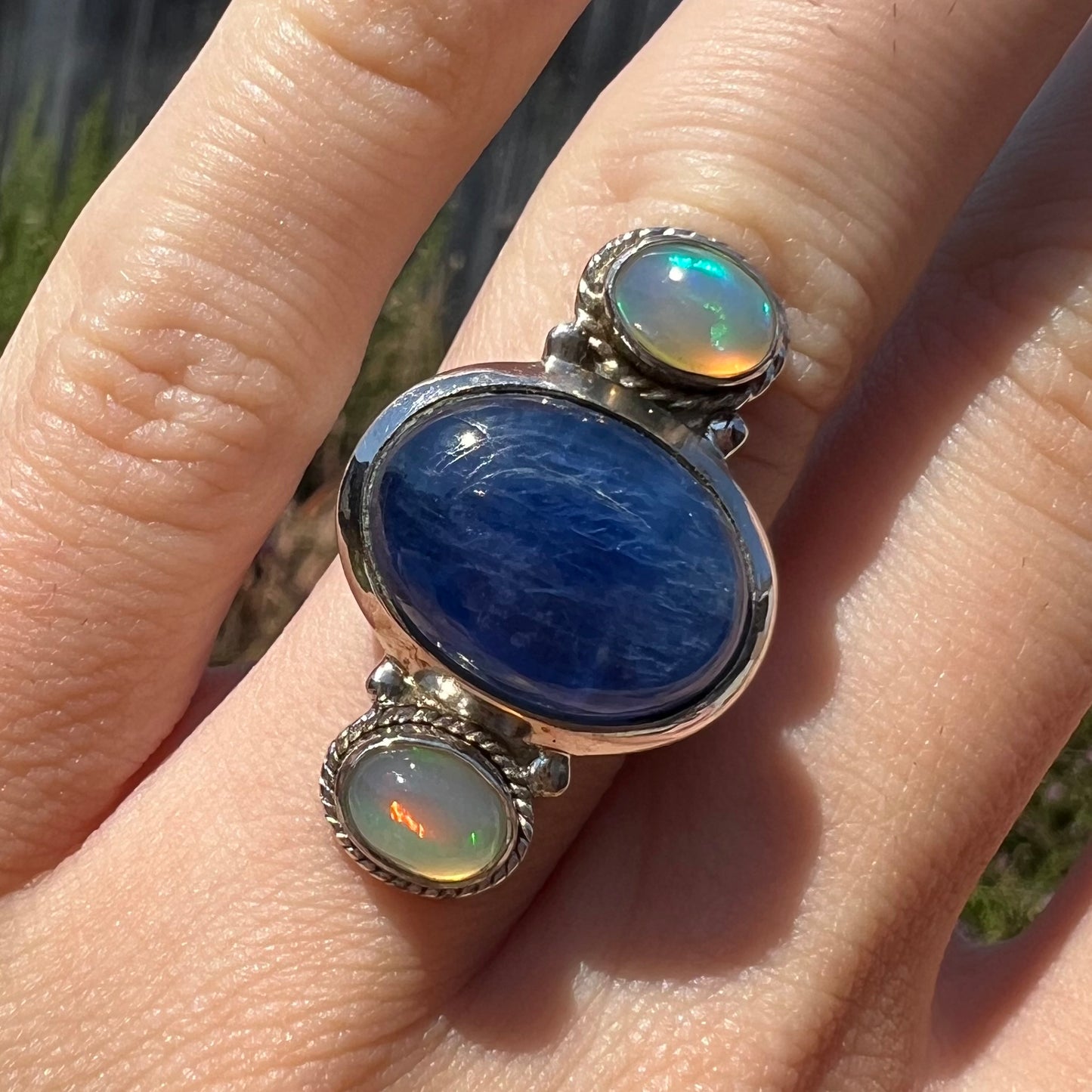 Front view close-up of a blue kyanite and opal ring worn on a finger in the sunlight.
