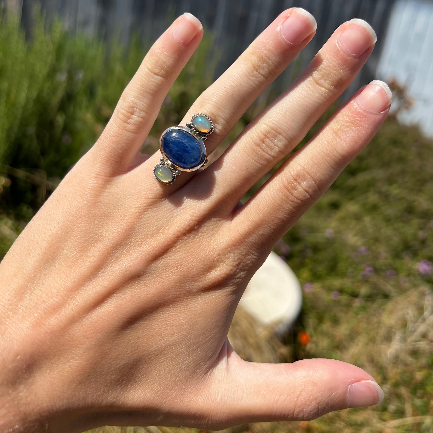 A blue kyanite and opal ring worn on a hand in the sunlight against a blurred outdoor backdrop.