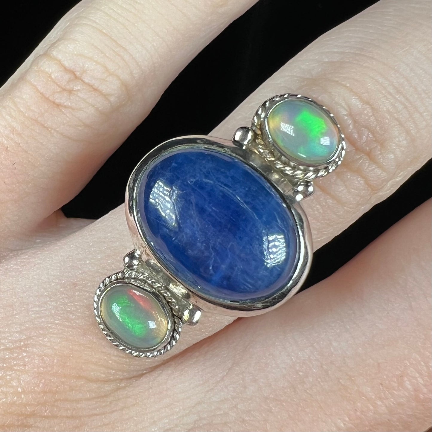 Close-up of a blue kyanite and opal ring worn on a finger against black background.