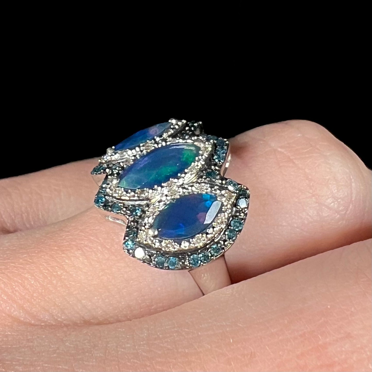 Angled view of a blue opal and diamond ring worn on a finger against a dark backdrop.
