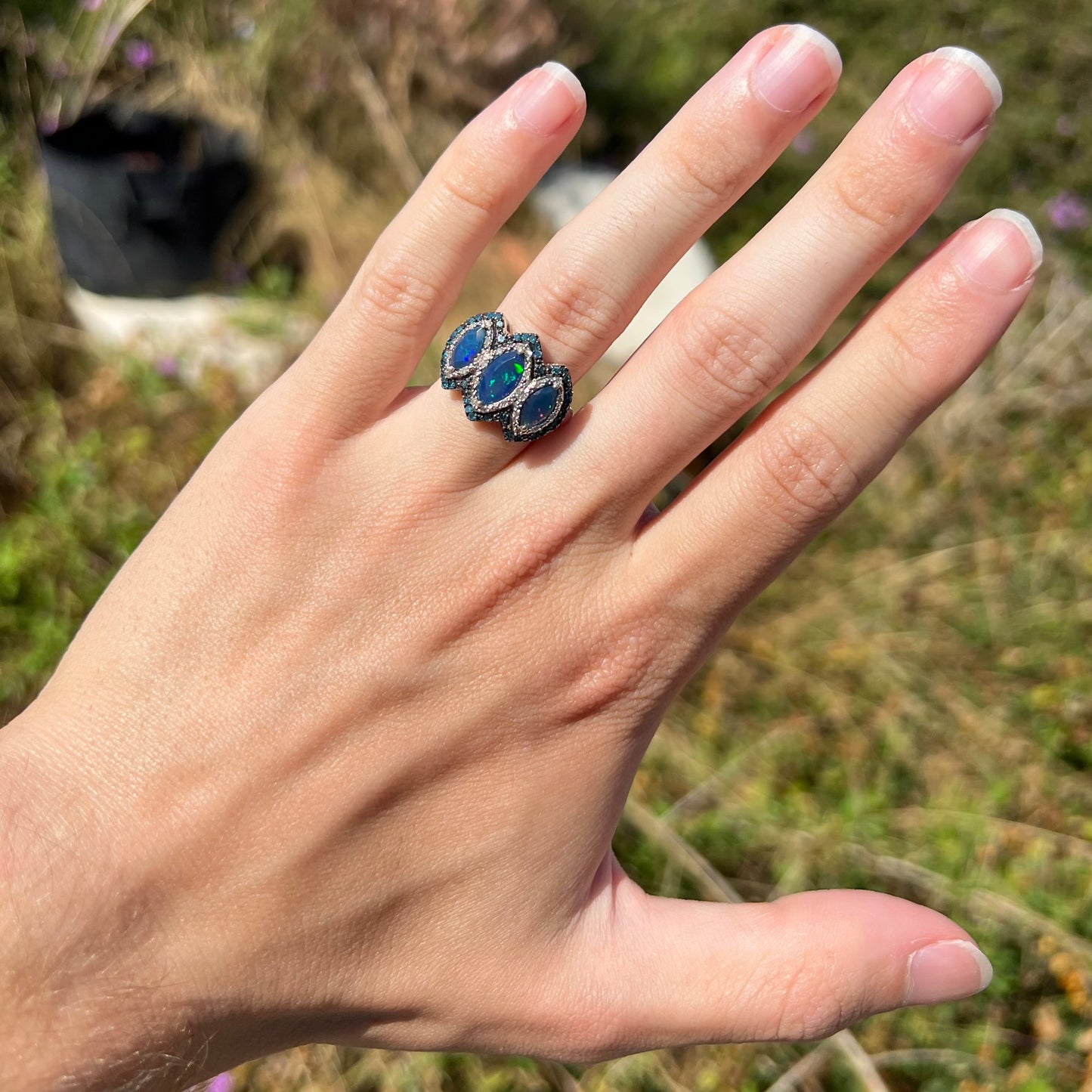 A marquise cut three-stone blue opal and diamond ring worn on a hand in natural sunlight.