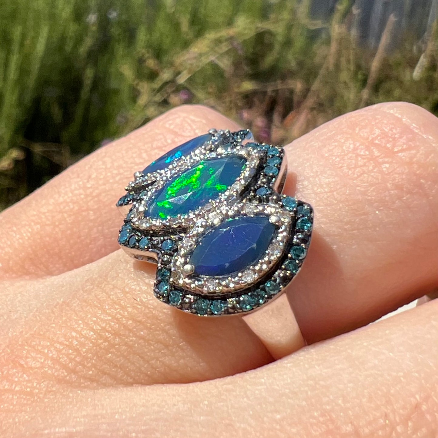 A three-stone marquise cut blue opal and diamond ring worn on a finger in sunlight at an angle. One opal has green fire.