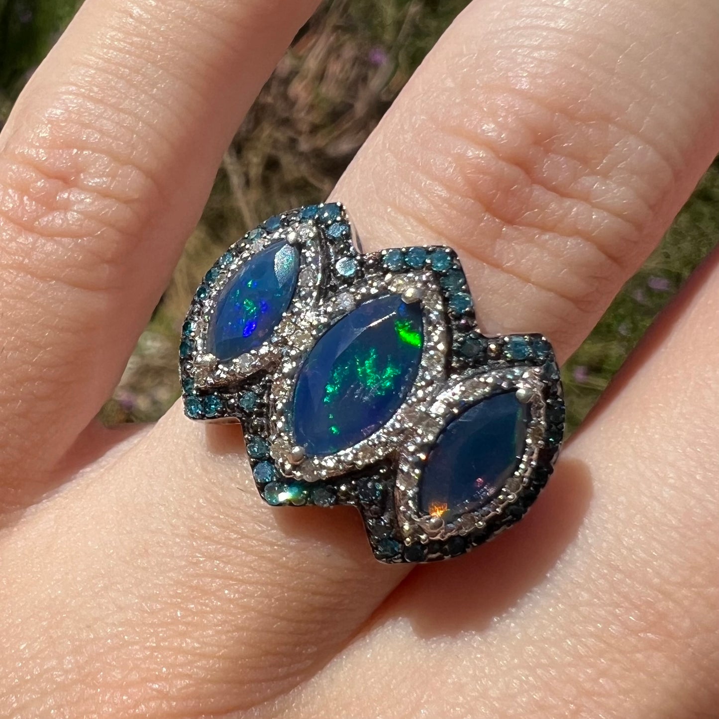 Close-up front view of a three-stone blue opal and diamond ring worn on a finger in the sunlight.