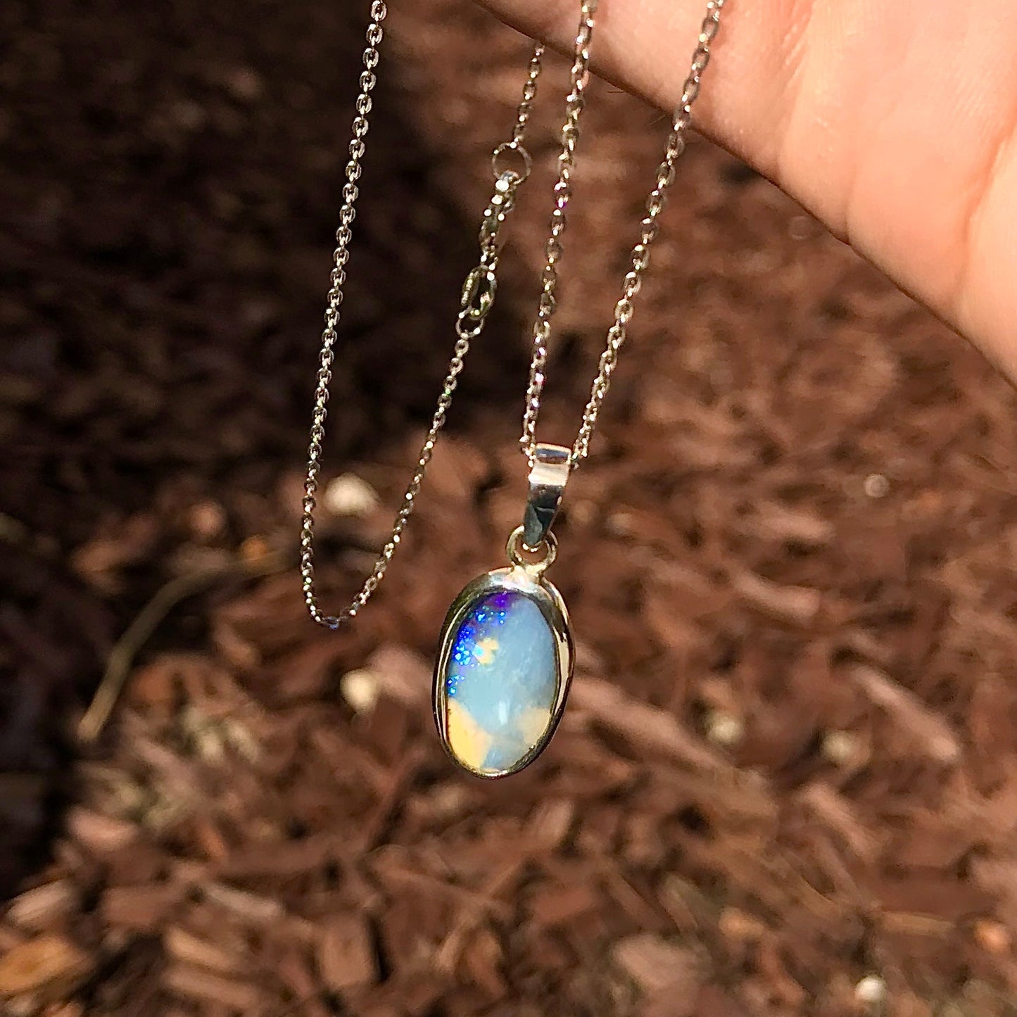 A photo of a Queensland Australian blue boulder opal with tan matrix set into a sterling silver bezel pendant.  The opal resembles a beach with tan sand matrix and a blue ocean of color.  The pendant is on a sterling silver cable chain and against a brown woodchip backdrop.