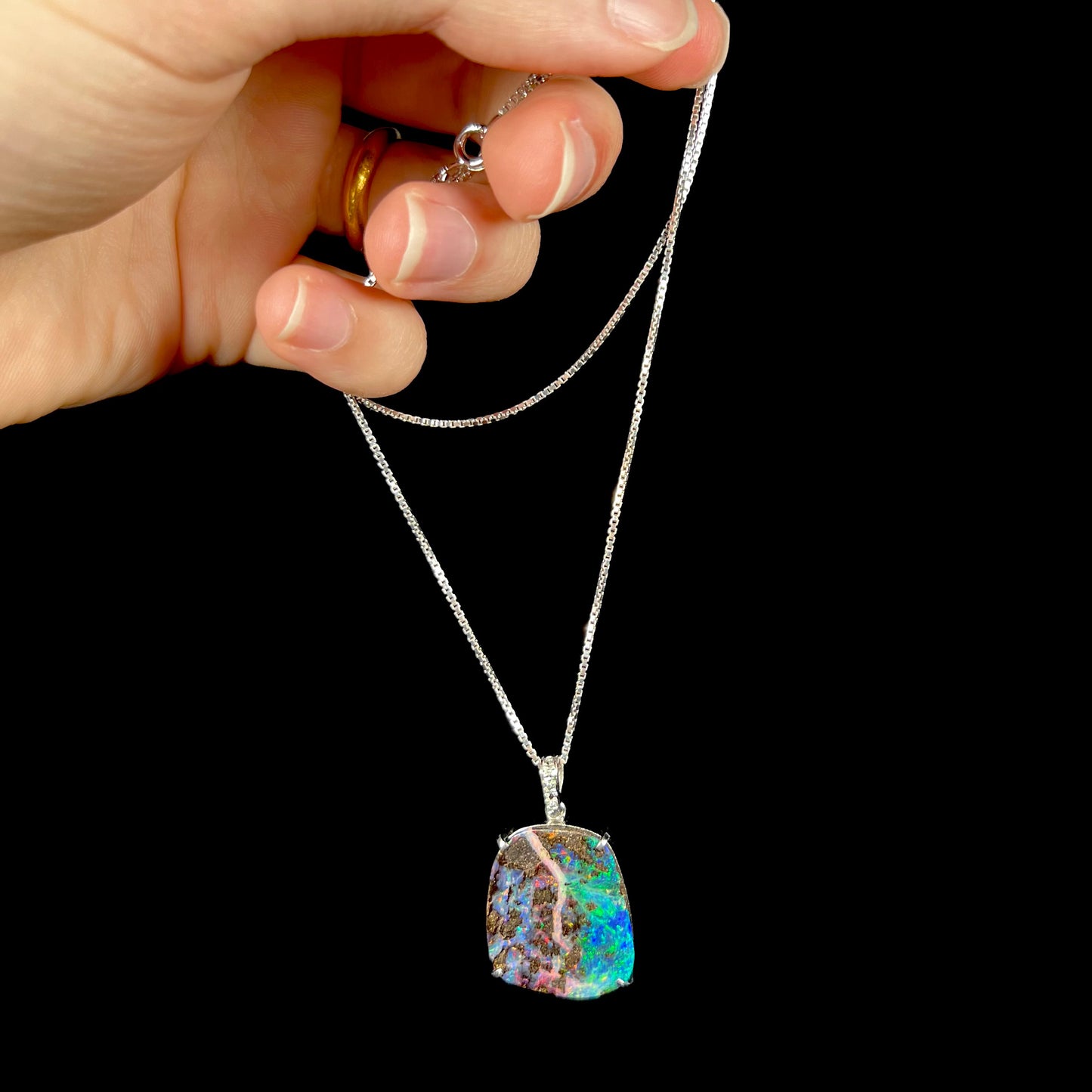 Platinum boulder opal necklace being held by the chain against a black background, showcasing its natural fire and matrix pattern.