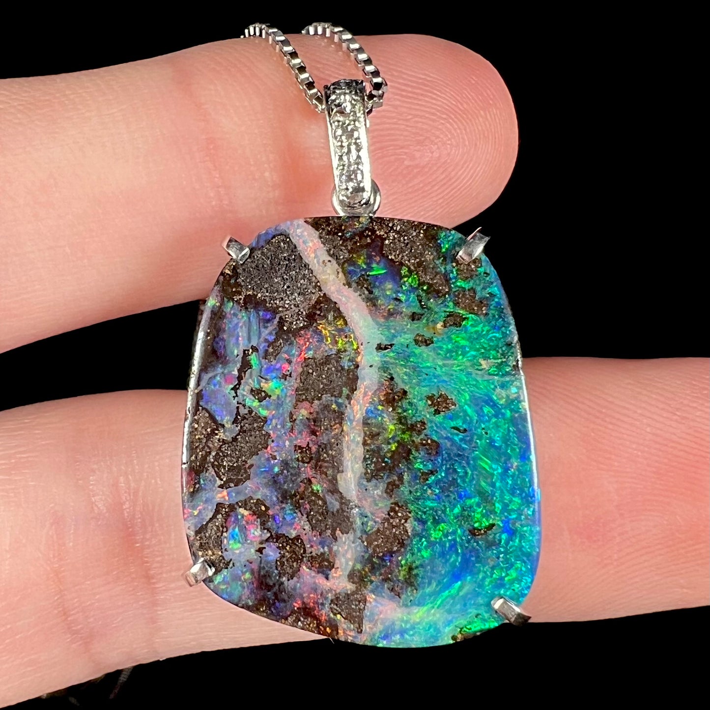 Macro view of a platinum boulder opal pendant resting on fingers, showcasing bright flashes of green, blue, and red.