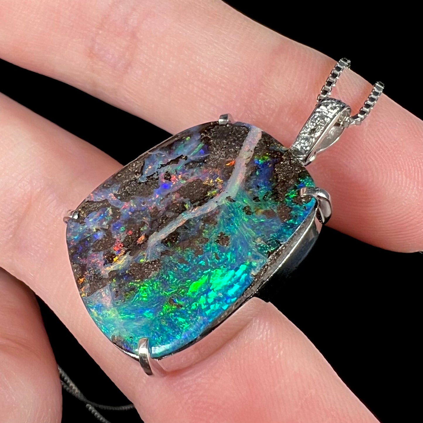 Macro angled shot of a platinum boulder opal pendant, with bright green and blue flashes reflecting from the opal’s surface.