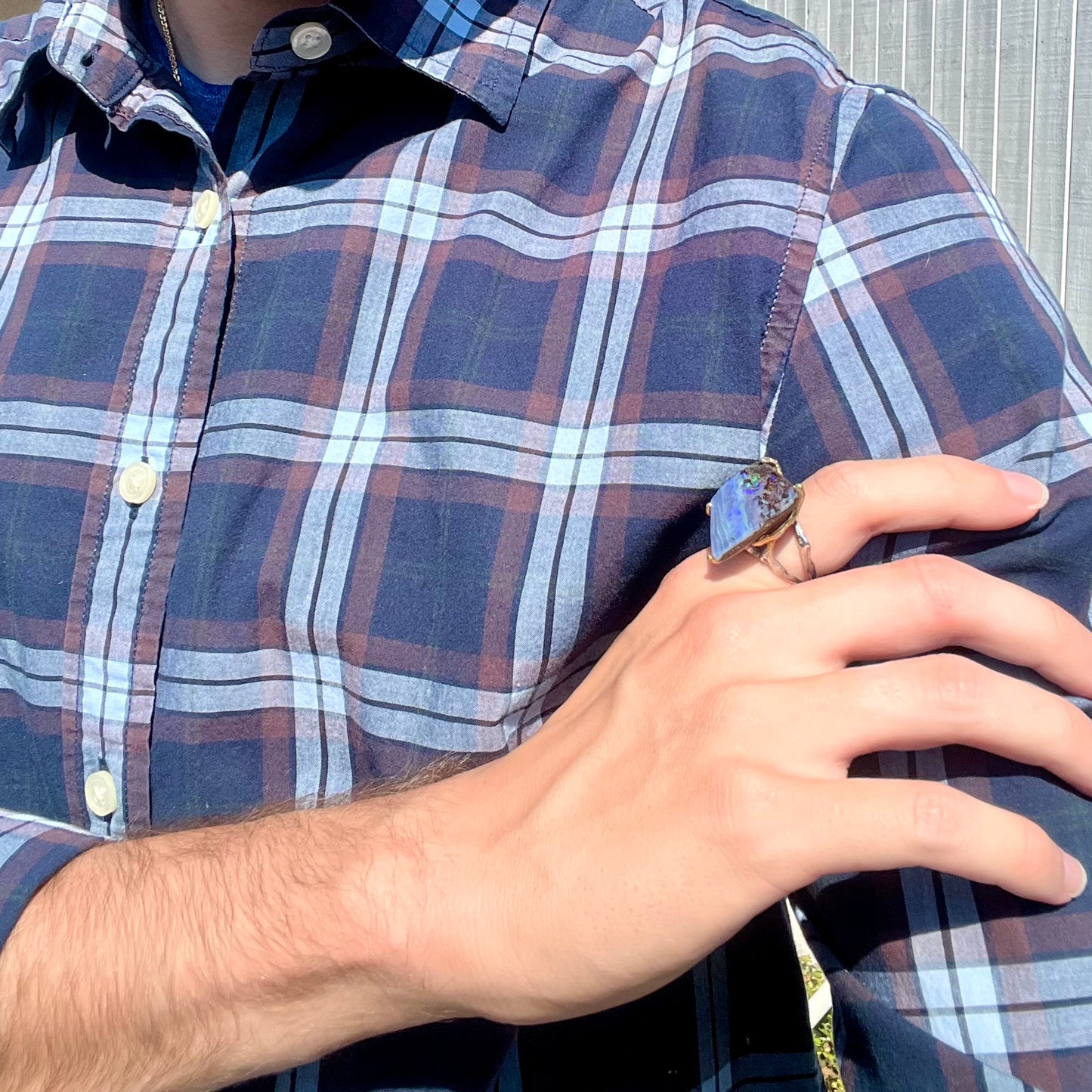 A person in blue plaid resting their hand on their shoulder, wearing a large Australian boulder opal ring.