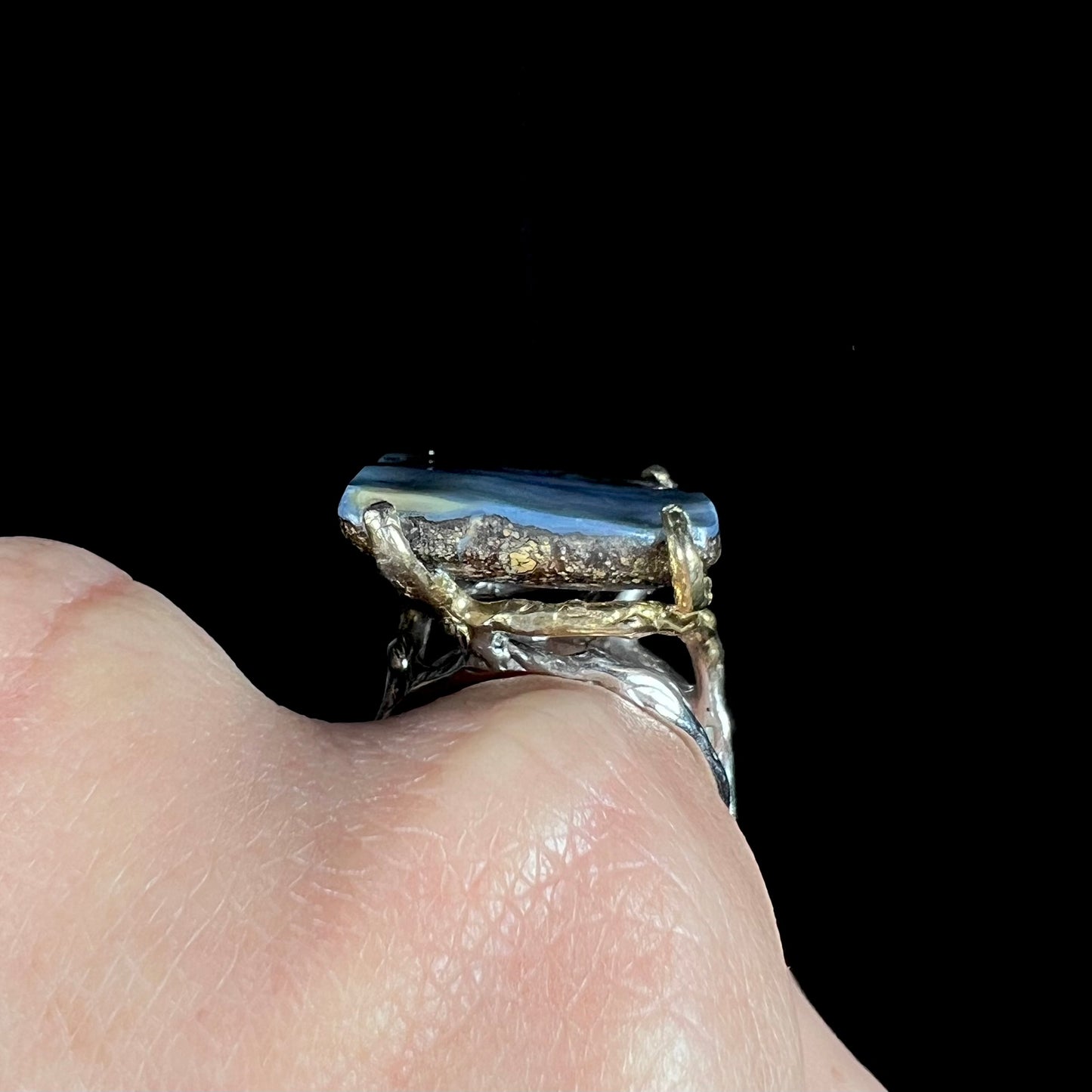 The side gallery view of a large boulder opal ring, worn on a hand, shown against a black backdrop.
