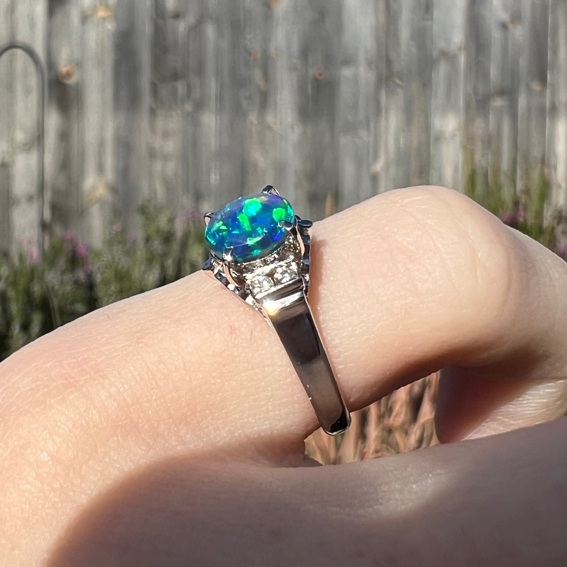 Bright blue and green colors are seen along the edge of a petite Lightning Ridge black opal and diamond ring worn on a pinkie finger.