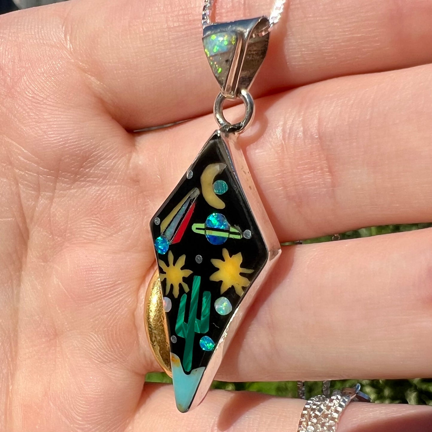 Silver opal and onyx inlay pendant with comet, moon, and star designs held in hand, shown from angle in natural sunlight.