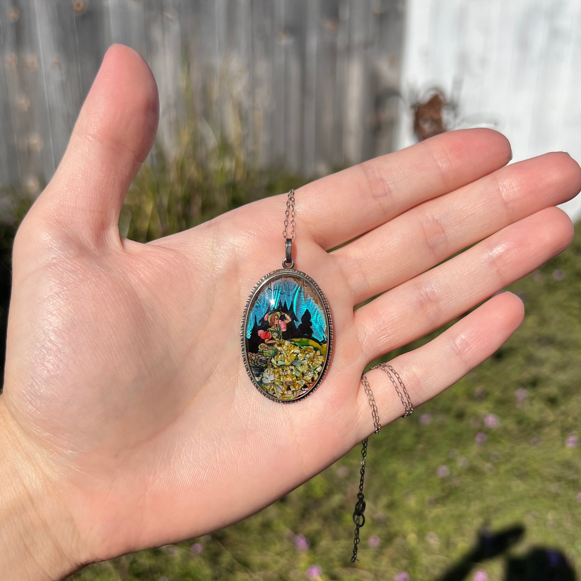 A crushed opal and butterfly wing necklace depicting the image of a woman, shown in a hand in sunlight.