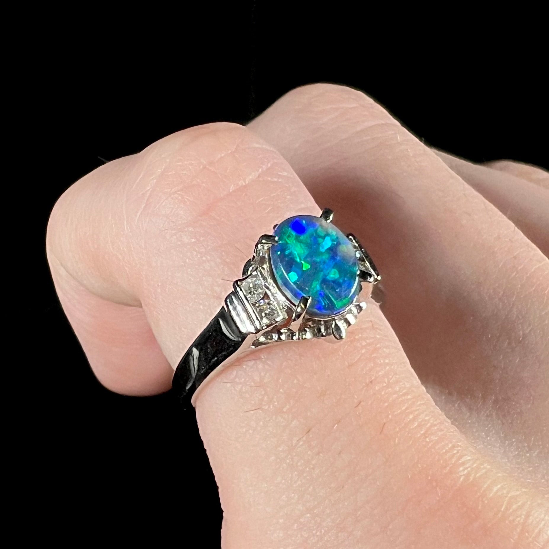 A dainty platinum black opal and diamond engagement ring worn on a pinkie finger.