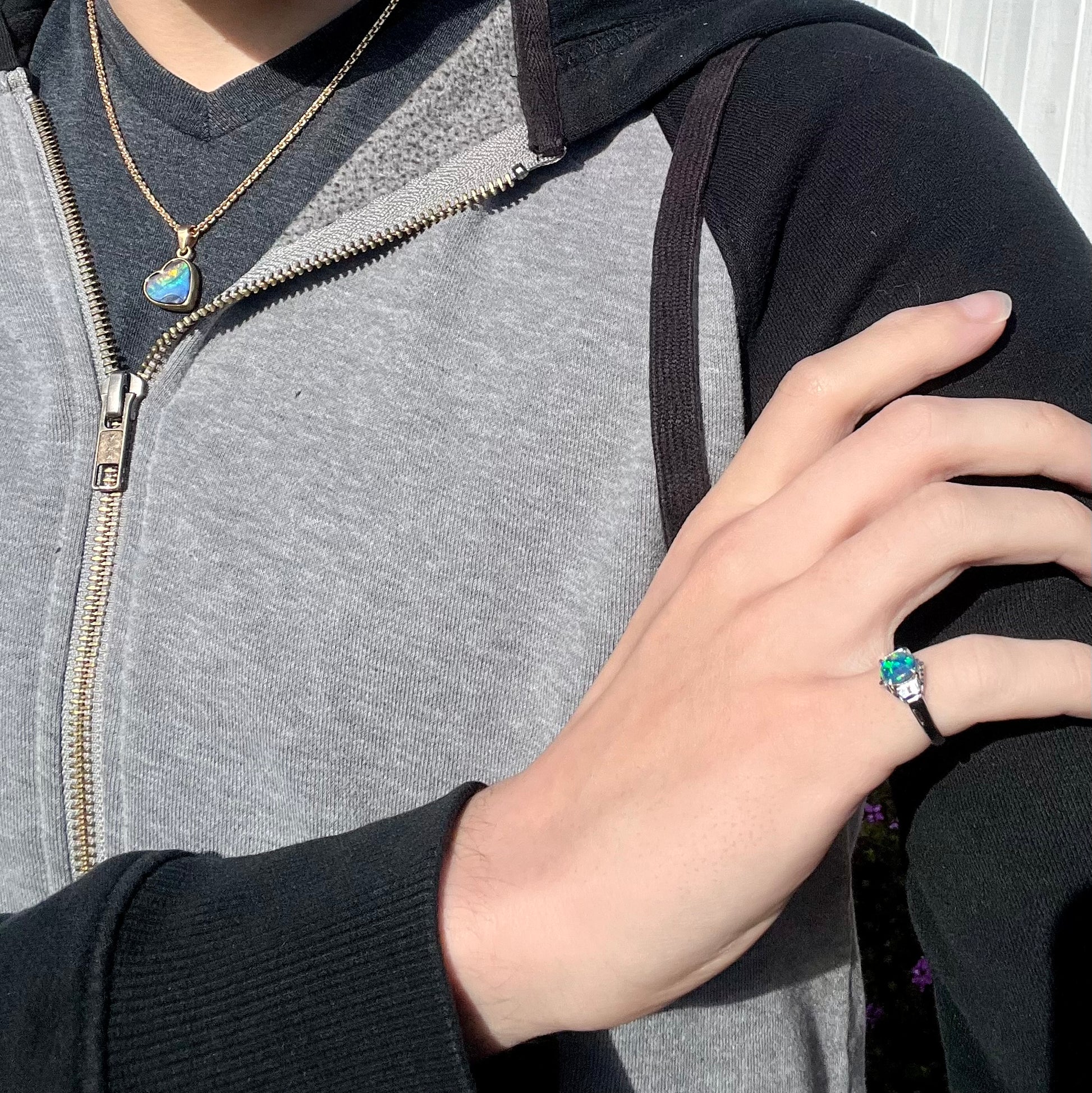 A model's hand wearing a dainty black opal engagement ring in the sunlight.  The opal's bright green colors are visible.