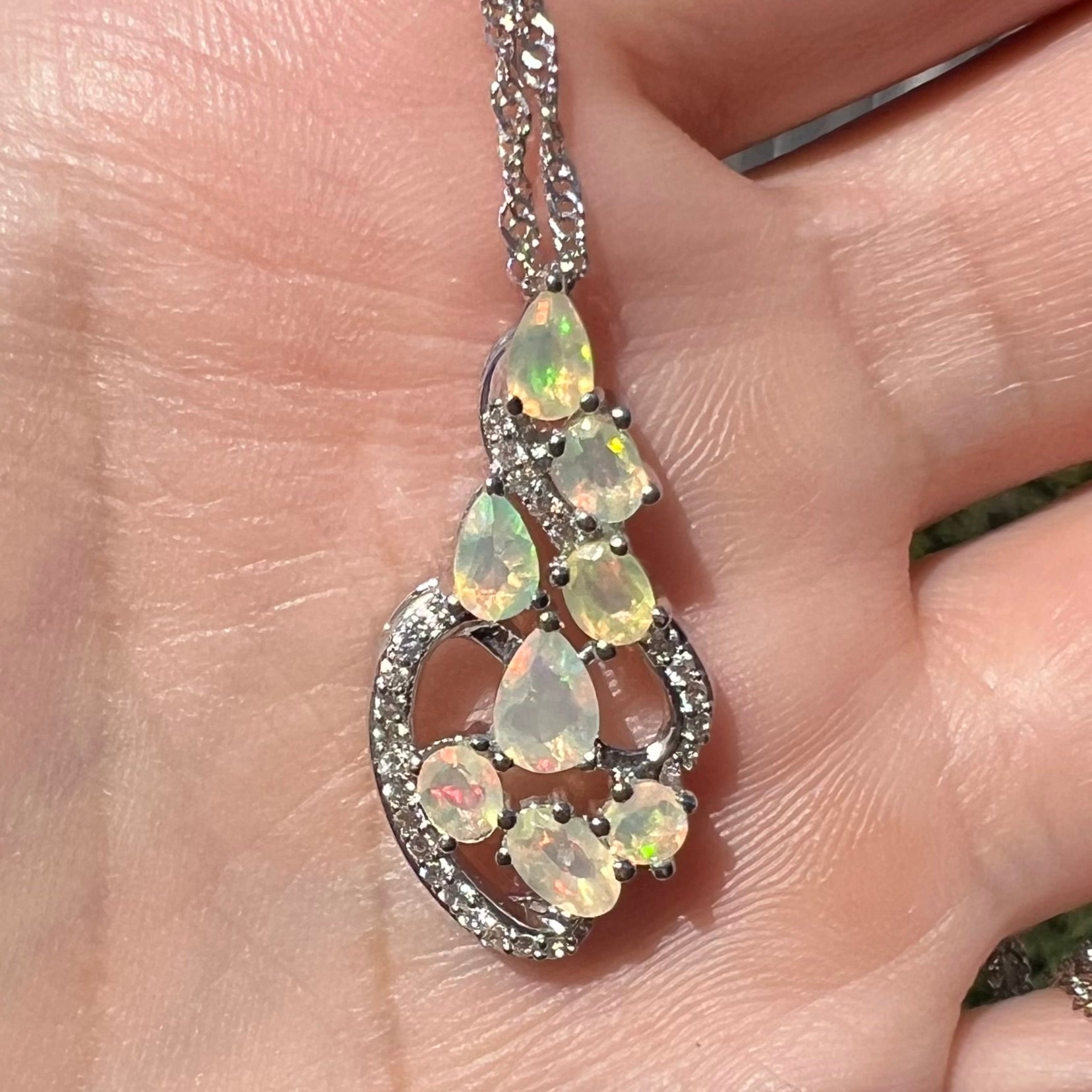A silver opal and CZ necklace with an elegant design, held by a hand and shown close-up in the sunlight.