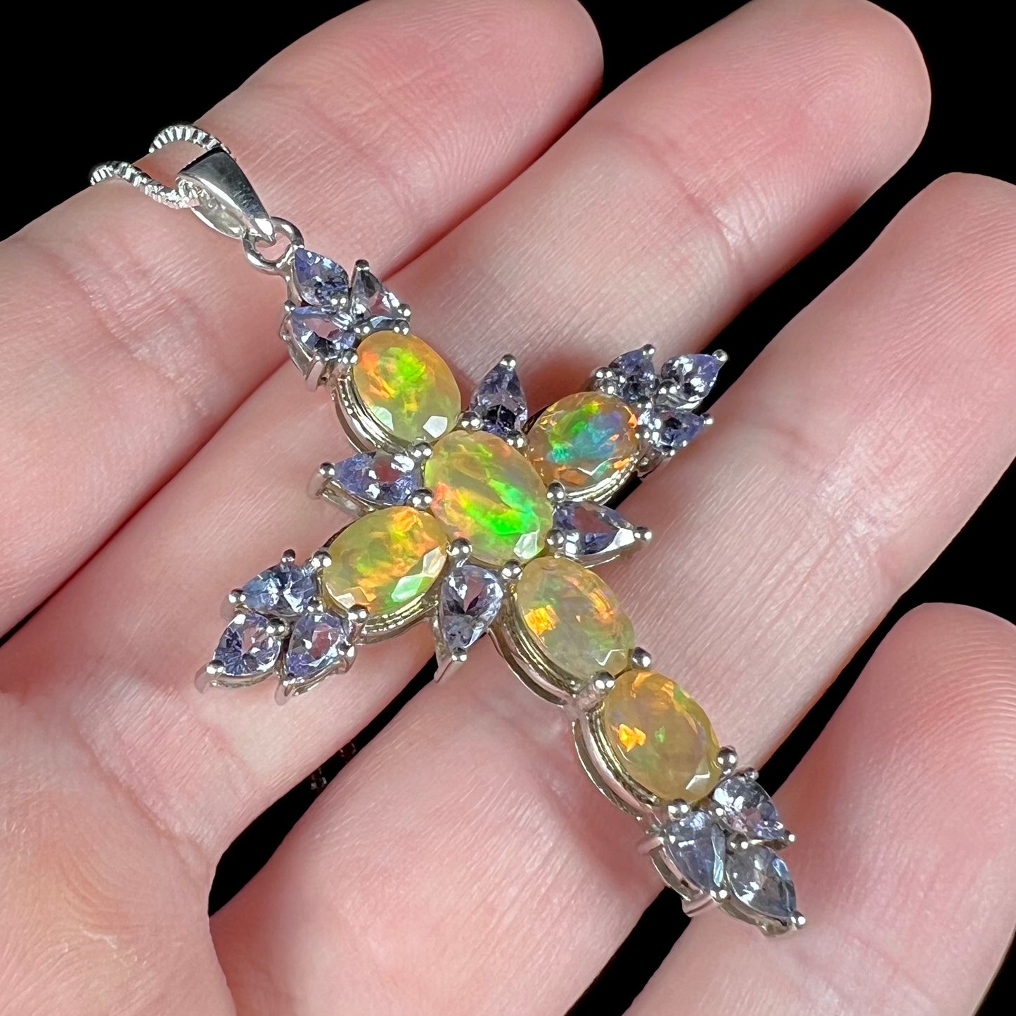 Angled close-up view of a hand holding an Ethiopian fire opal cross necklace with tanzanite accents.