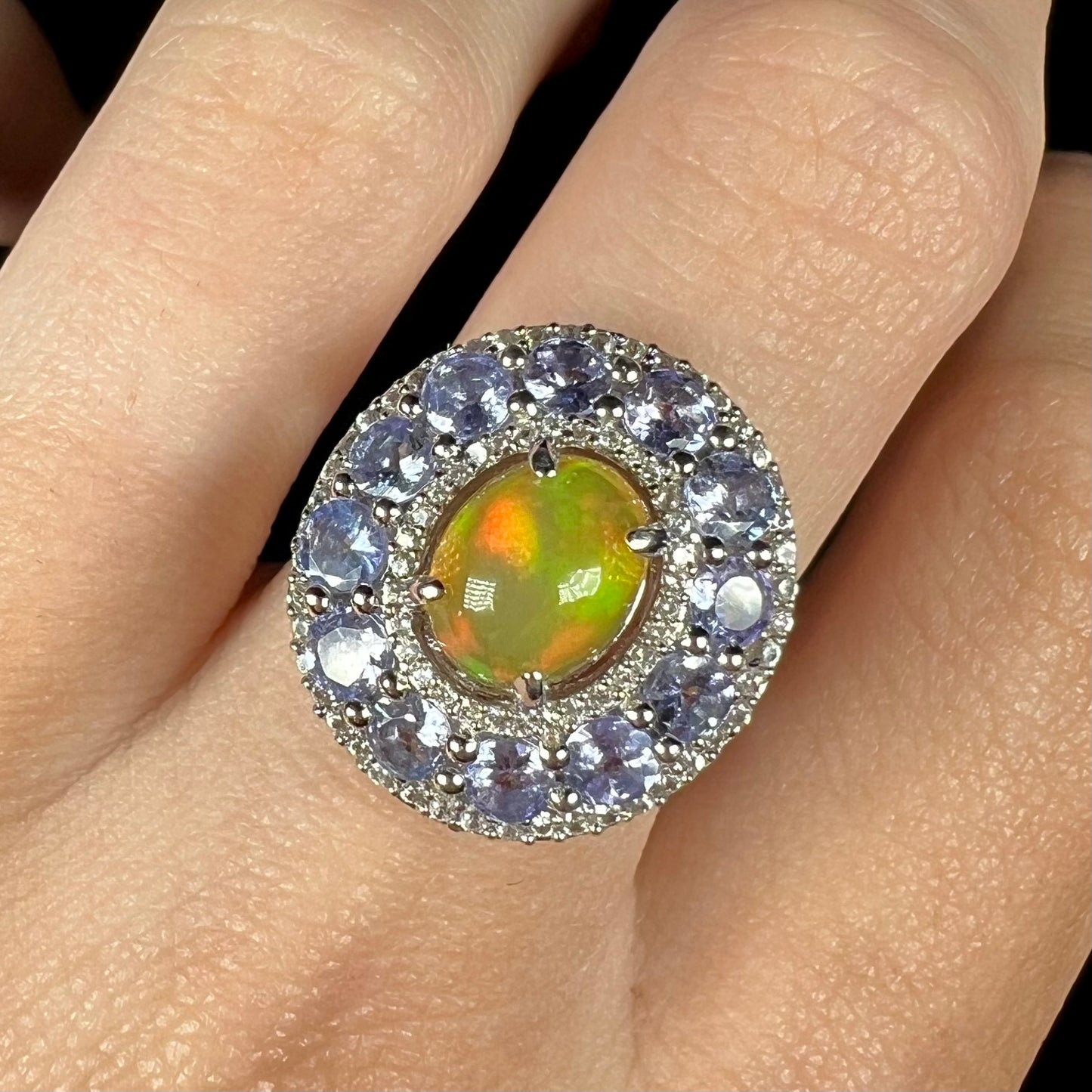 Close up of hand wearing Ethiopian fire opal ring with tanzanite and white zircon halos against black background.