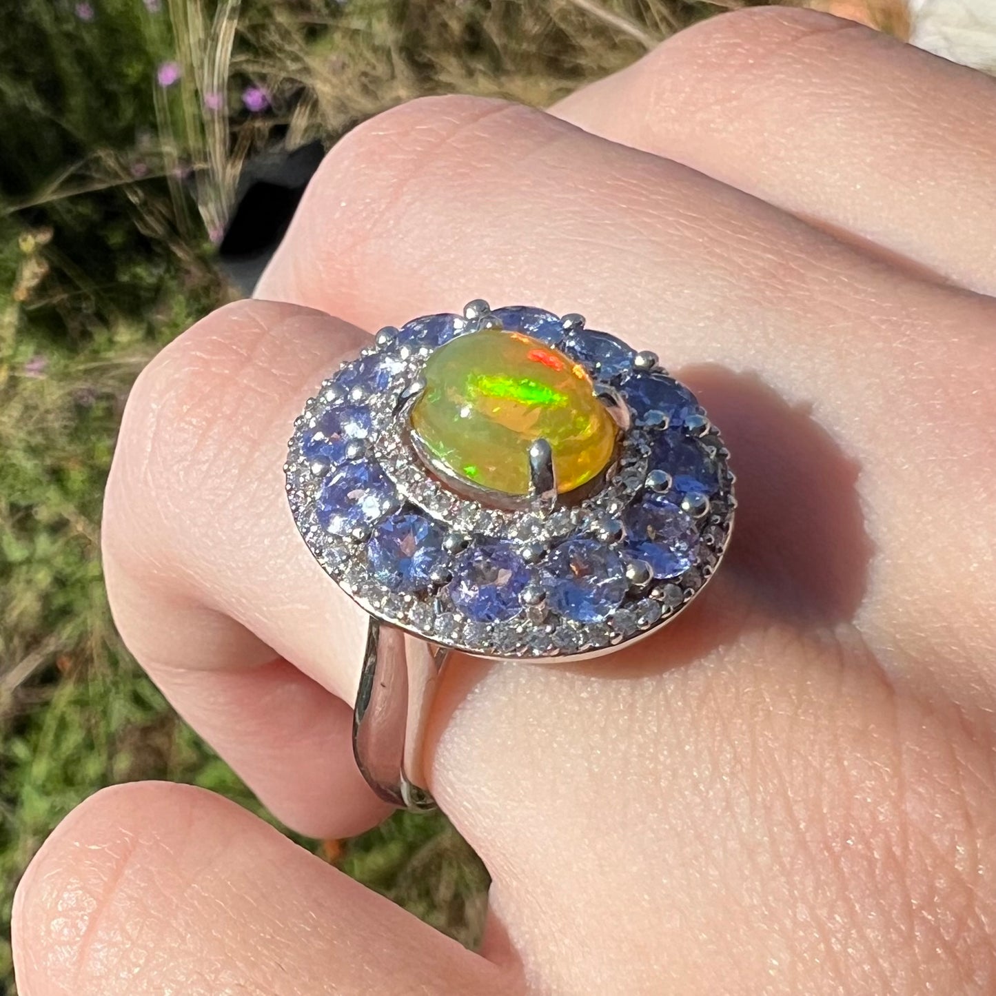 Close up angled shot of hand wearing Ethiopian fire opal and tanzanite halo ring in natural sunlight.