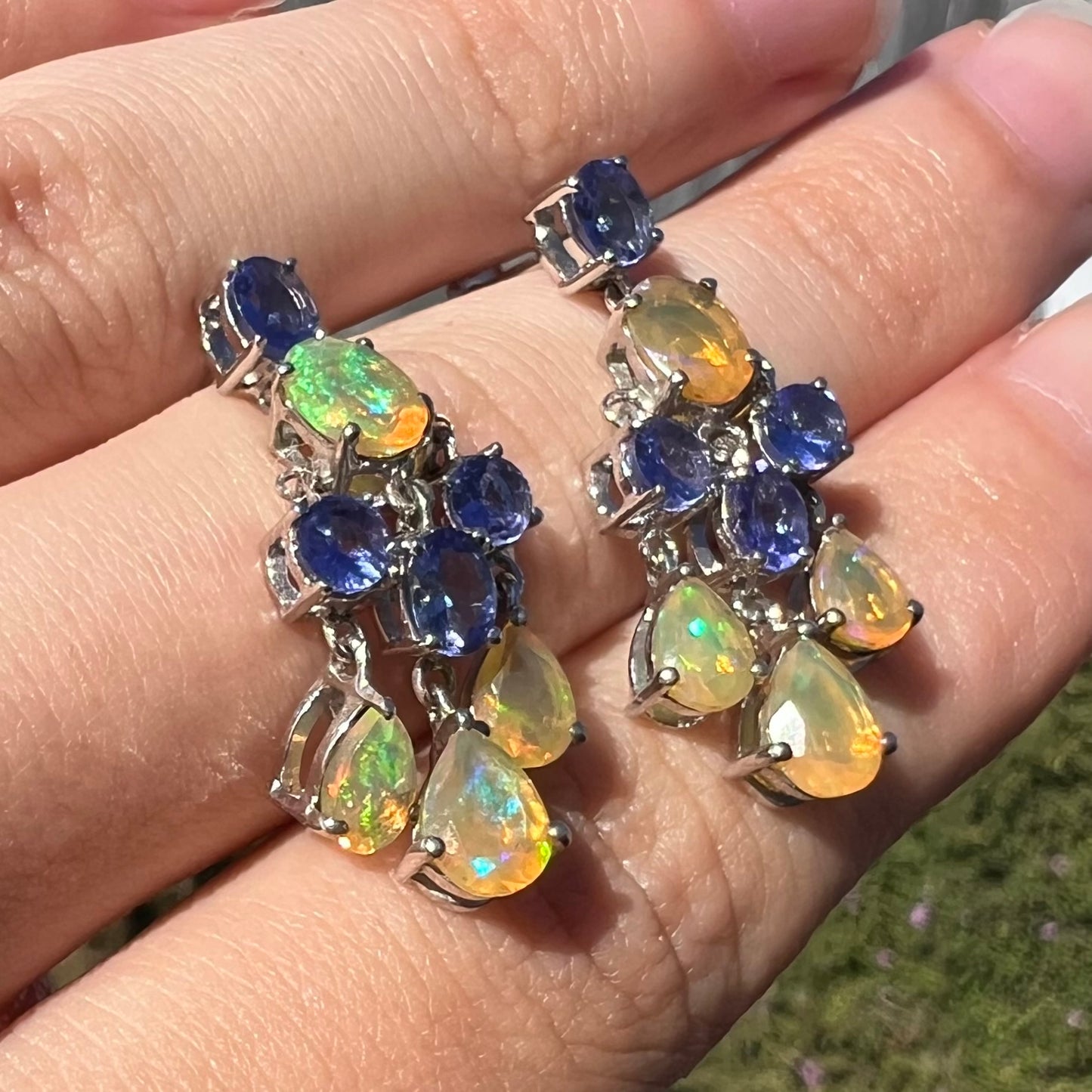 A pair of dangling sterling silver Ethiopian opal and tanzanite chandelier earrings, shown at an angle in sunlight.