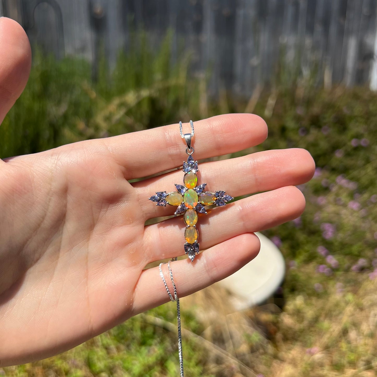 An Ethiopian opal cross necklace with tanzanite accents, held in a hand at a distance in the sunlight.