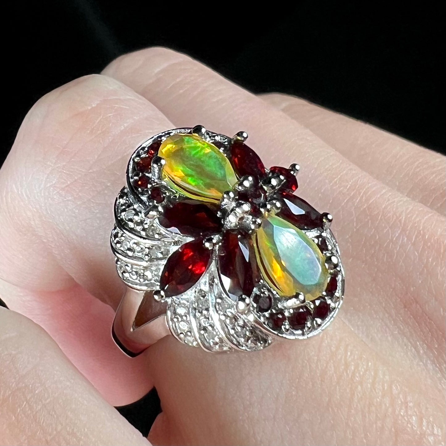 A sterling silver Ethiopian opal and garnet infinity-design ring with diamond accents, worn on a finger at an angle.