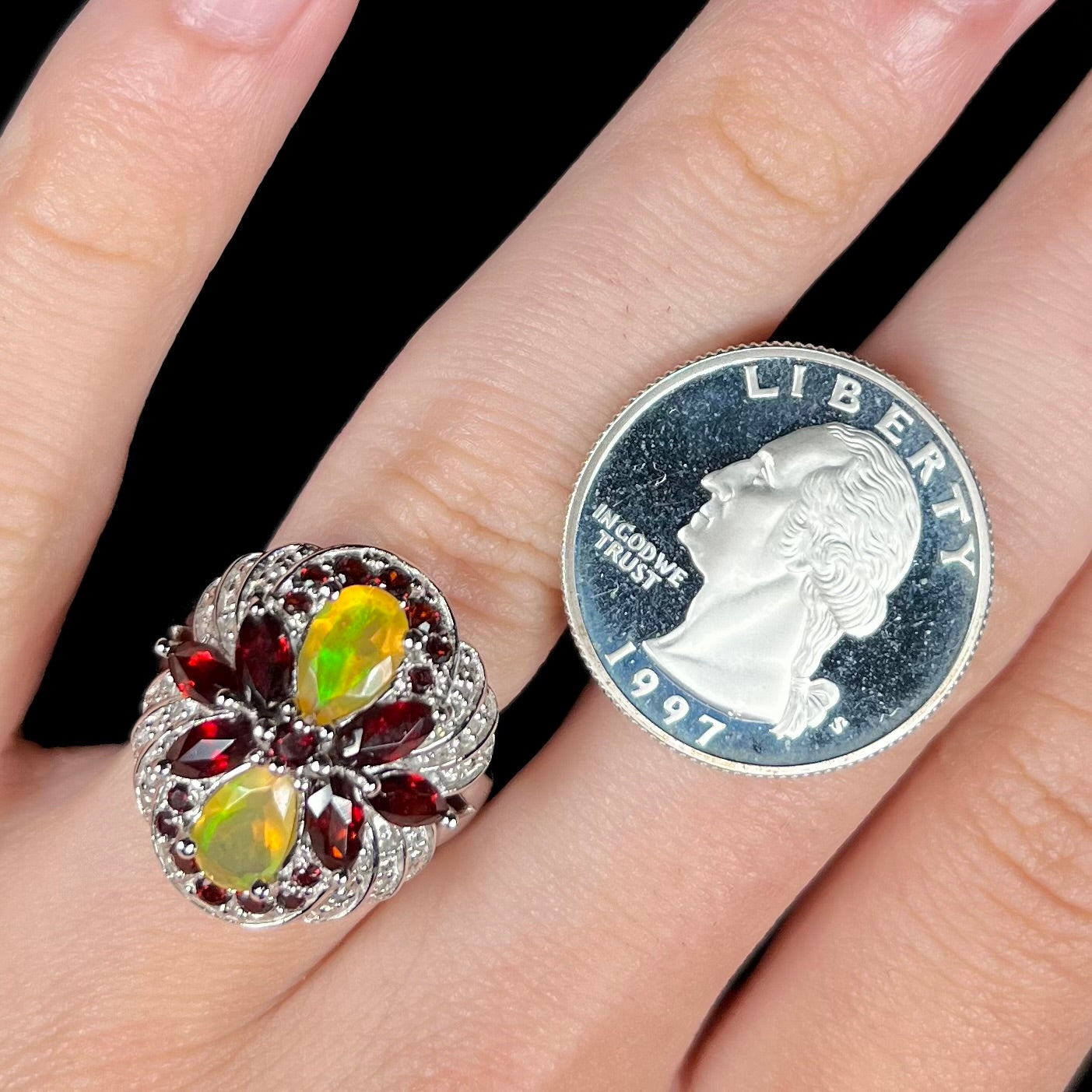 A sterling silver Ethiopian opal and garnet infinity-design ring with diamond accents worn on a finger next to a quarter coin.