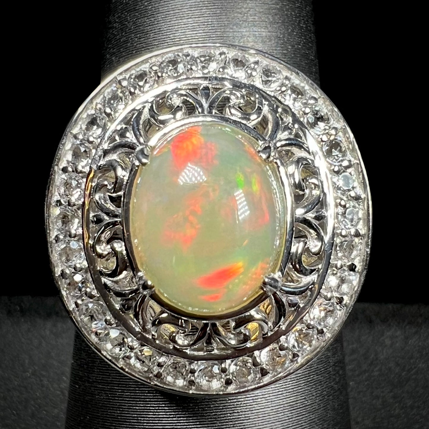 Front view of Ethiopian opal ring with double halo of silver filigree and white topaz against black background.