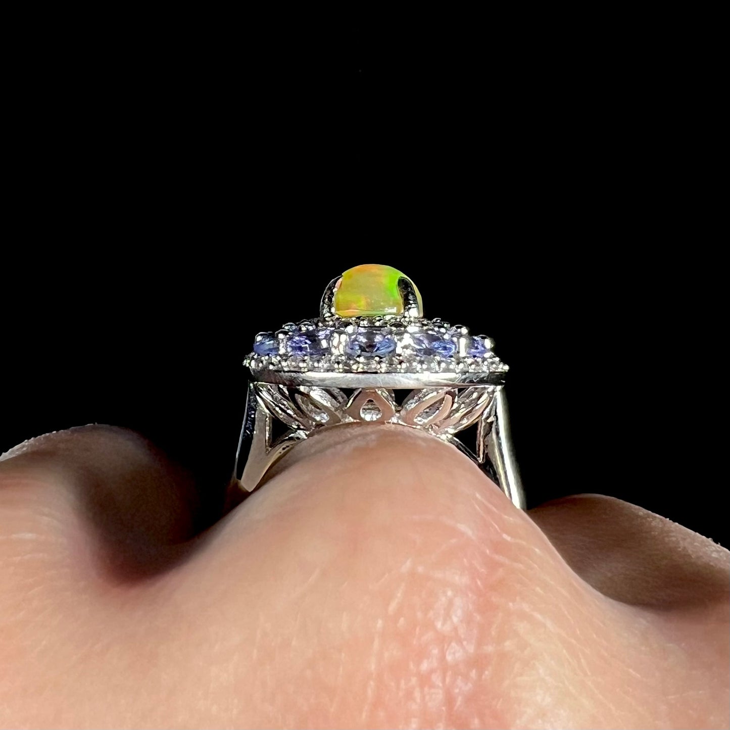 Macro gallery view of silver Ethiopian opal halo ring worn on a hand against black background.