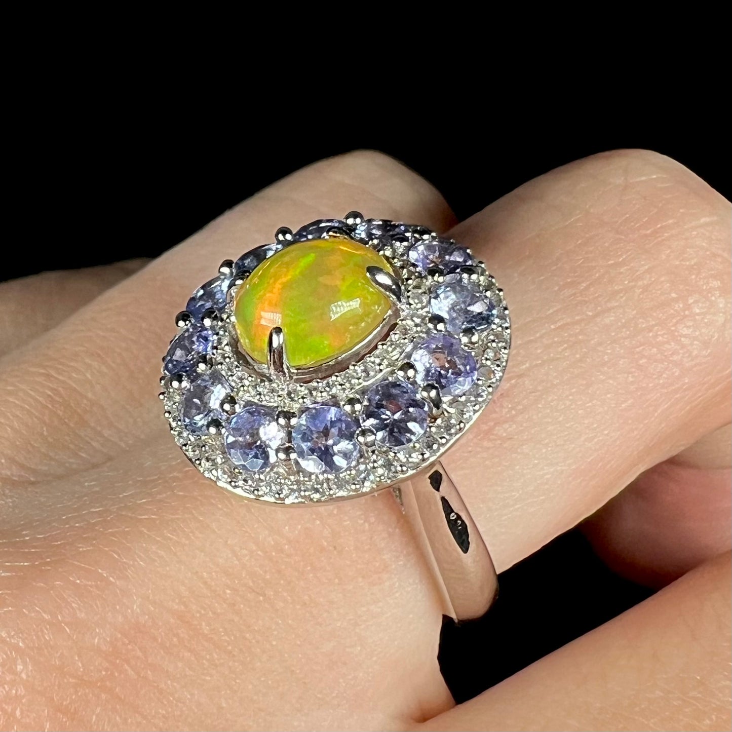 Angled close up of Ethiopian opal halo ring worn on a hand against black background showing tanzanites.