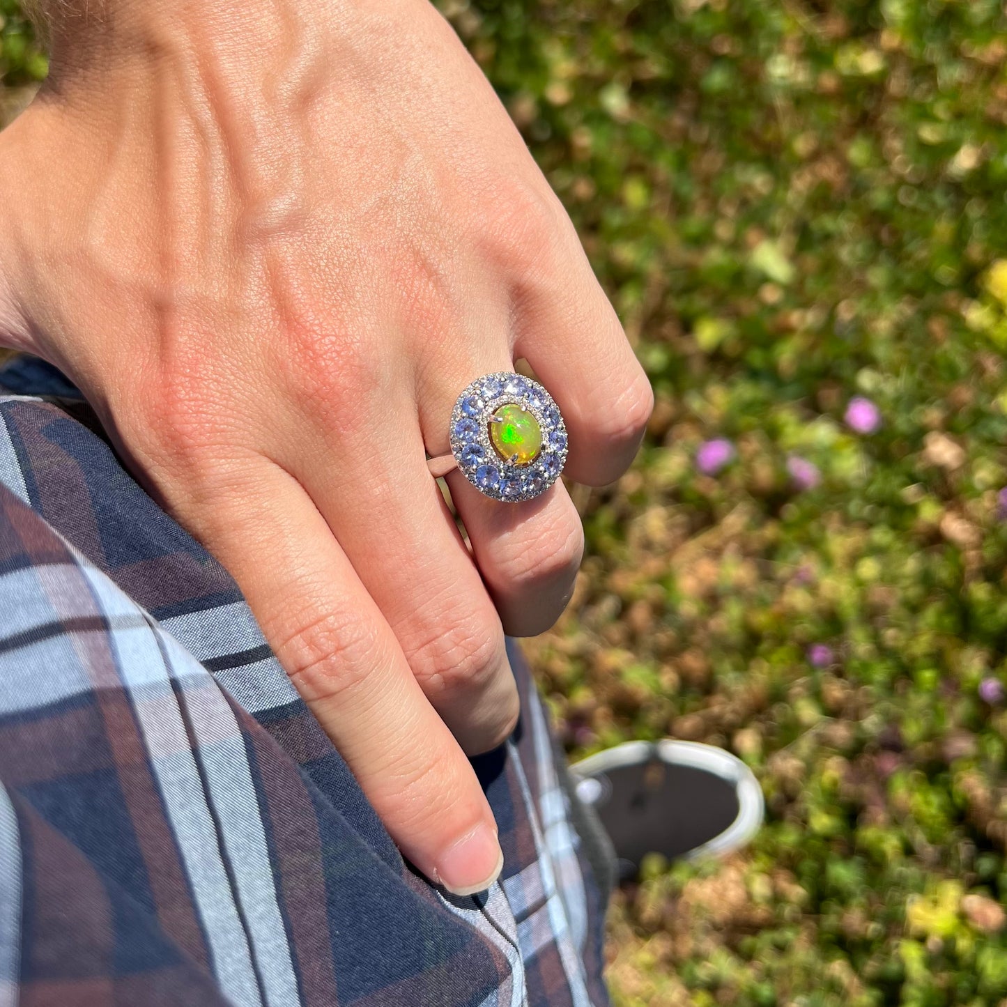 Person's hand on hip wearing Ethiopian opal and tanzanite halo ring in sunlight. Green color play is visible in the opal.