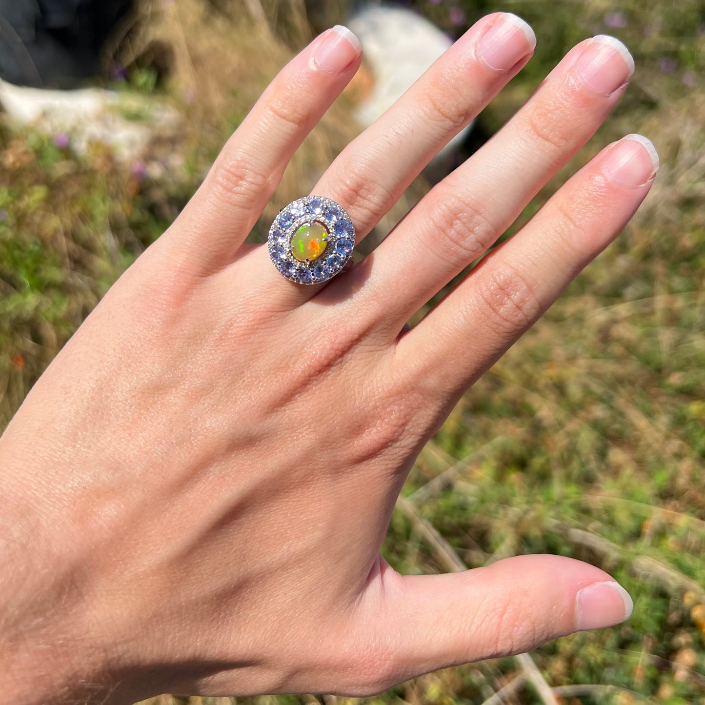 Hand wearing Ethiopian opal and tanzanite halo ring in sunlight. Red and green colors seen in opal.