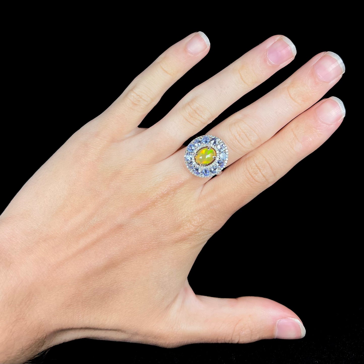 Hand wearing Ethiopian opal ring with tanzanite halo against black background.