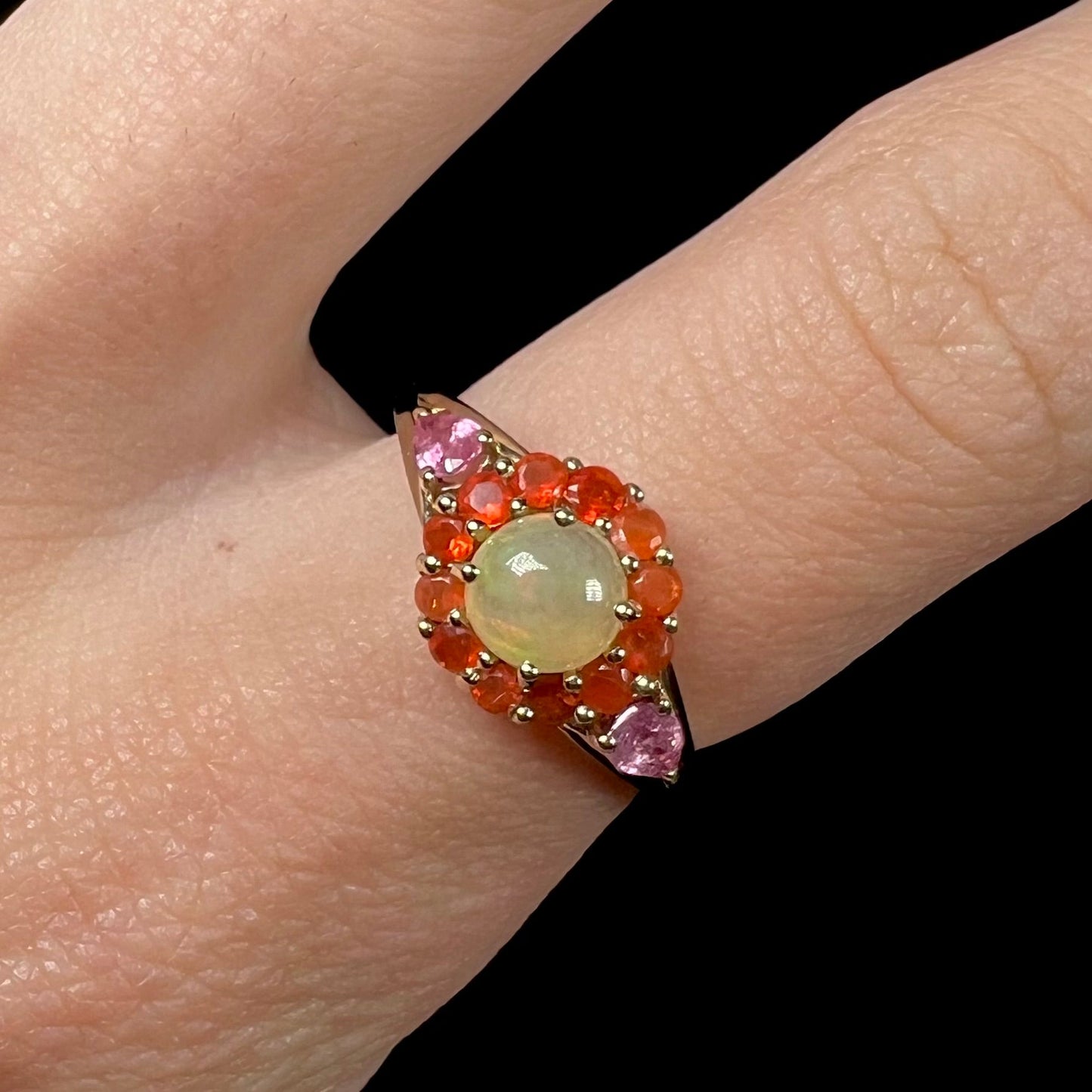 A yellow gold ring mounted with a round Ethiopian opal in a halo of faceted Mexican fire opals. Two pear shaped pink tourmalines accent the sides of the ring.